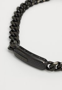 Bracelet en chaîne noire en métal, avec un panneau texturé gravé de "GUESS" et une rangée d'accents noirs pour le détail.