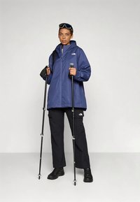 Mėlyna nepralaidi vandeniui striukė su gaubtu, juodos krovinių kelnės ir juodi žygių batai. Rankose du žygių lazdos, ant striukės ir kelnių baltas logotipas.