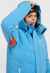 Blauwe pufferjas met een hoge kraag, voorzien van een ritssluiting aan de voorkant, oranje accenten en elastische manchetten voor comfort en warmte.