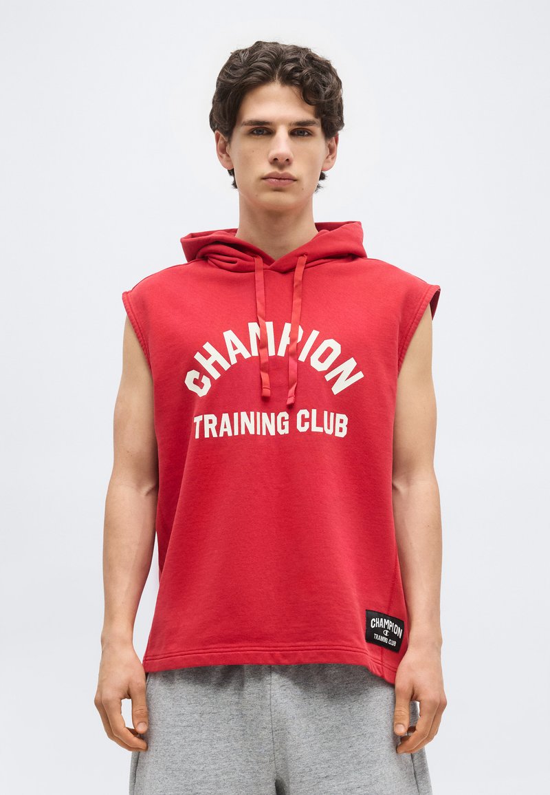 Young man wearing red sleeveless hoodie with "Champion Training Club" text and gray sweatpants, standing against a plain light background.