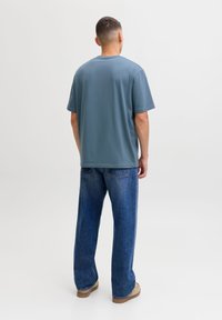 Blue, short-sleeve t-shirt paired with relaxed-fit, medium-blue denim jeans. The outfit is completed with beige shoes. Back view shown.
