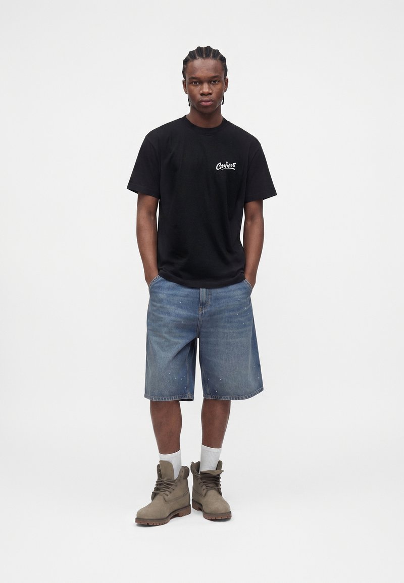 Ung mand stående med hænderne i lommerne iført sort Carhartt t-shirt, blå denimshorts, hvide sokker og beige snørestøvler.