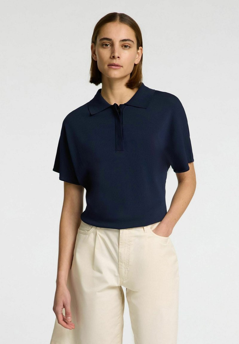 Mørkeblå polo-shirt med kort, struktureret ærmedesign og knapstolpe, parret med cremefarvede højtaljede bukser; glat tekstur.