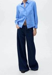 Person in einem hellblauen Hemd mit Knopfleiste und weit geschnittenen dunkelblauen Jeans, der mit einer Hand in der Tasche vor einem schlichten Hintergrund steht.