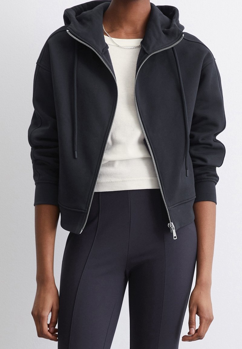 Svart zip-up-hoodie med fodrad huva, kenguruficka och ribbade muddar. Bärs över en ljus topp och kombineras med mörka, figurnära byxor.