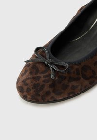 Bailarina de ante marrón con borde negro y detalle de lazo en la parte frontal, con un patrón de leopardo en la parte superior. Forro interior suave.