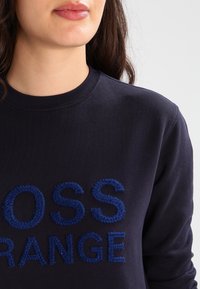 Marinblå bomullströja med broderad blå text "CROSS ORANGE" på framsidan, ribbad rund halsringning och mjuk textur.