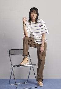 Randig vit och lila t-shirt, bruna cargobyxor och beige högskurna sneakers. Modellen poserar medan hen lutar sig mot en kromad hopfällbar stol.