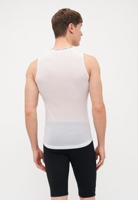 Chemise sans manches blanche en maille avec un ourlet arrondi et des accents noirs. Ajustement près du corps ; portée avec un short de cyclisme noir, montrant la vue arrière.