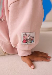 La mano de un niño descansando en el suelo, vistiendo una manga rosa con un parche que muestra a tres cerdos de dibujos animados abrazándose.