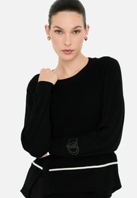 Maglione nero lavorato a maglia con maniche lunghe, caratterizzato da una texture a coste, una striscia bianca di accento all'orlo e un logo ricamato sulla manica.