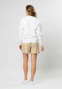 Baltas krekls ar garām piedurknēm, apaļu kakla izgriezumu un trikotāžas aprocēm, saskaņots ar bēšiem šortiem, skatīts no aizmugures. Baltas kedas noslēdz kopējo izskatu.