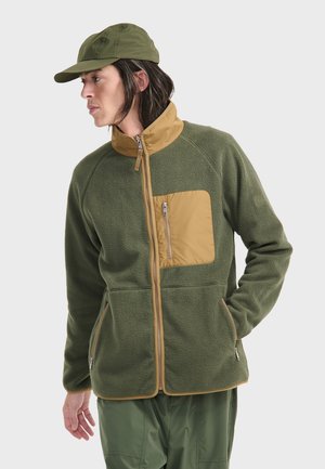 Veste en polaire verte avec une poche poitrine marron, fermeture éclair sur le devant et col montant. Tissu texturé avec des poches latérales et des finitions contrastées.