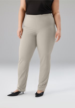 Frau trägt beige enge Hose, schwarzes ärmelloses Oberteil und schwarze Kitten-Heel-Schuhe und steht vor einem einfarbigen grauen Hintergrund.