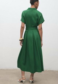 Green, knee-length dress with a collar, short sleeves, and a belted waist. Features a pleated skirt and smooth texture. Worn with pale sandals.