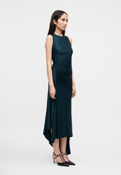 Naine seisab tumerohelises varrukatuta voltides midi-kleit koos asümmeetrilise äärega ning mustade teravate varvastega kontsakingades, millel on pahkluu rihmad.