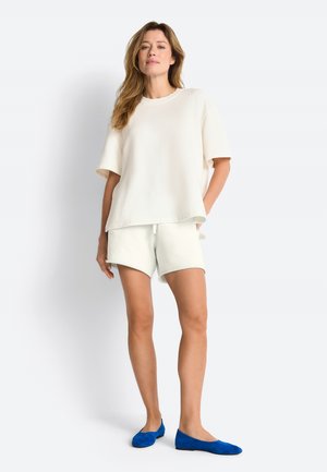 Frau trägt ein lockeres weißes kurzärmeliges Oberteil und passende Shorts, steht mit einer Hand in der Tasche und trägt leuchtend blaue Ballerinas.