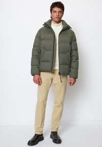 Olive green puffer jacket with horizontal quilting, a hood, and a front zipper. Paired with beige pants and black boots, featuring a slim fit.