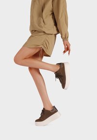 Gambe in sneaker marroni con suole bianche a piattaforma, indossando una gonna beige arricciata e una giacca coordinata su uno sfondo semplice.