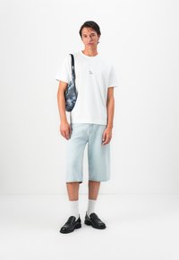 Homem a usar uma t-shirt gráfica branca, calções de ganga azul claro, meias brancas, sapatos mocassins pretos e a carregar uma mala de ombro azul com padrão, contra um fundo branco.