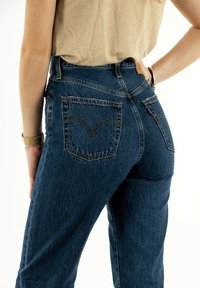 Femme portant un jean en denim bleu taille haute et un top beige, avec une main reposant sur sa hanche et l'autre détendue à ses côtés.
