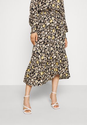 Vrouw draagt een zwarte midi-rok met bloemenprint in geel en wit, gecombineerd met witte sandalen met hoge hak en bandjes.