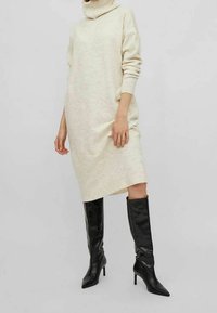 Abito lavorato a maglia color crema con collo a imbuto, maniche lunghe e vestibilità rilassata, abbinato a stivali neri in pelle lucida alti fino al ginocchio con tacco.