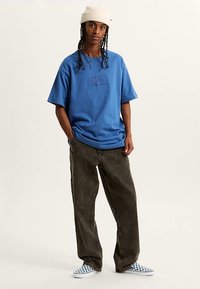 Blå bomullst-shirt med präglad logotyp, kombinerad med lösa bruna jeans och rutig slip-on-skor. Modellen bär en krämfärgad mössa.