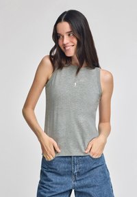 Graues ärmelloses Oberteil aus weichem Stoff, mit rundem Halsausschnitt und einem kleinen Logodetail. Kombiniert mit hochgeschnittenen Jeans.