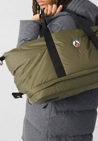 Persona che indossa un giubbotto grigio di tipo puffer, mentre tiene una grande borsa a tracolla verde oliva con manici neri e un logo rotondo JOTT.