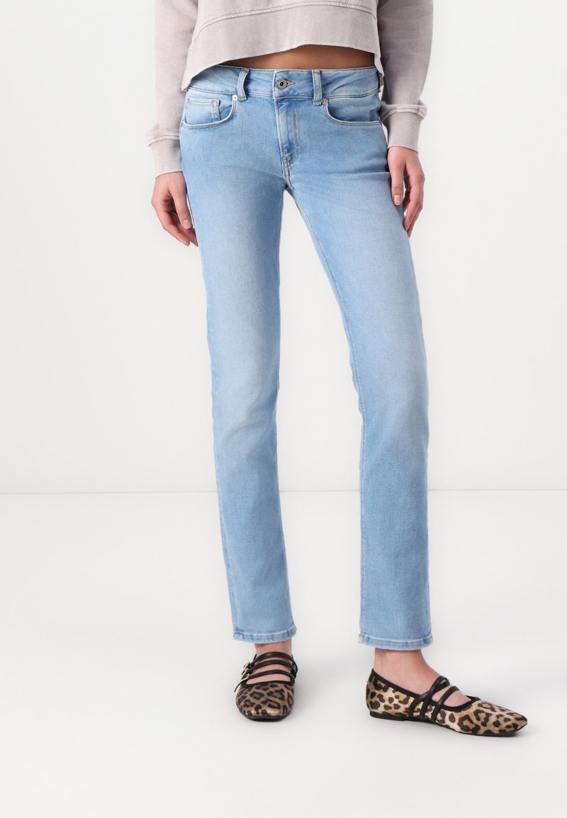 Personne portant un jean bleu clair à jambe droite, un sweat-shirt court gris clair et des ballerines à imprimé léopard avec des brides noires, debout sur un sol blanc.