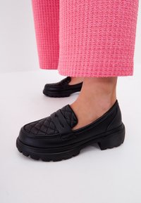 Zwarte gewatteerde instappers met een chunky zool, voorzien van een gladde afwerking en een klassiek banddetail, gecombineerd met roze gestructureerde wijde broeken.