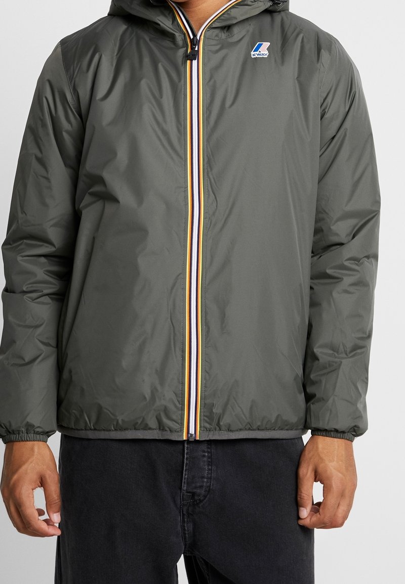 Homme portant une veste à capuche zippée vert olive avec une fermeture éclair rayée et le logo K-Way sur la poitrine, associée à un pantalon noir.