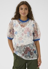 Frau, die ein transparentes Oberteil aus blumiger Spitze mit blauen Akzenten über einem einfarbigen blauen Shirt trägt, kombiniert mit dunkelgrünen Hosen, vor einem einfarbigen Hintergrund stehend.