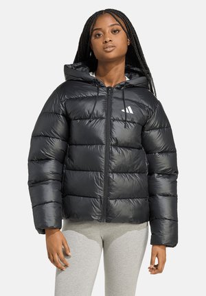Jeune femme aux longs cheveux tressés portant une doudoune noire à capuche et un pantalon gris clair, debout devant un fond uni.