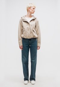 Άτομο με κοντά ξανθά μαλλιά, που φοράει μπεζ σακάκι, λευκό πουκάμισο, μπλε τζιν και λευκά παπούτσια, στέκεται και κοιτάζει προς τα δεξιά.