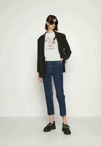 Μαύρο στιλιζαρισμένο σακάκι πάνω από λευκό Τ-shirt με γραφικό σχέδιο, συνδυασμένο με μπλε τζιν παντελόνι και μαύρες πλατφόρμες.