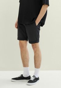 Czarne szorty z podwiniętymi nogawkami, wykonane z lekkiego materiału, w połączeniu z czarnymi sneakersami i białymi skarpetkami do kostek. Prosty design, brak wyraźnych wzorów.