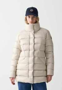 Chaqueta acolchada de color beige claro con cuello alto, diseño acolchado horizontal y diagonal, que presenta bolsillos laterales y un parche con logo en la manga.