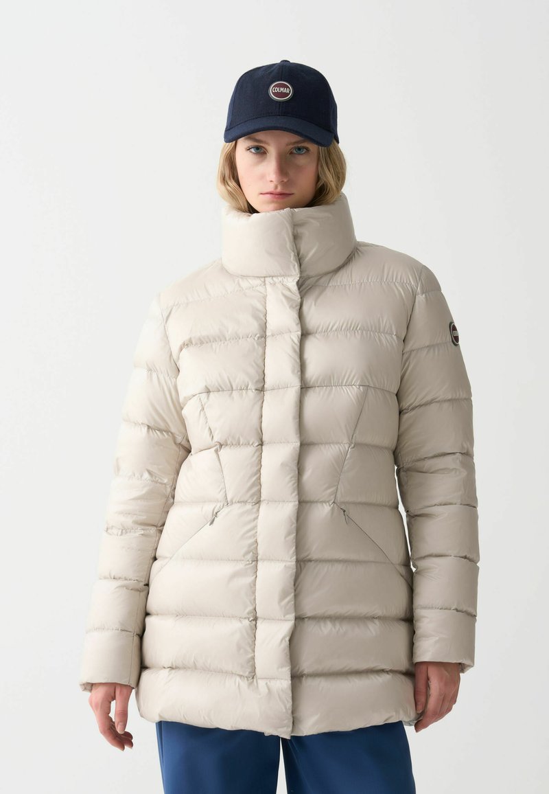 Chaqueta acolchada de color beige claro con cuello alto, diseño acolchado horizontal y diagonal, que presenta bolsillos laterales y un parche con logo en la manga.