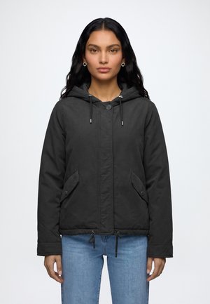 Femme aux longs cheveux noirs, portant une veste noire à capuche avec des poches avant et un jean bleu clair, debout devant un fond blanc.
