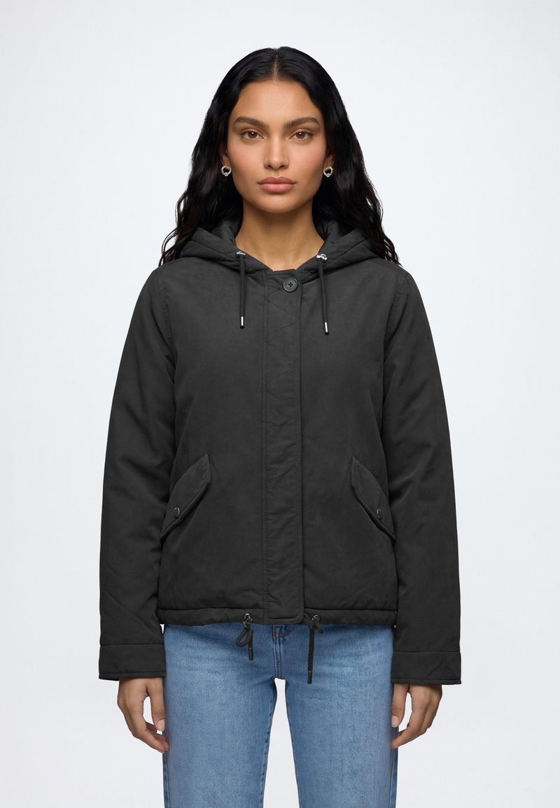 Femme aux longs cheveux noirs, portant une veste noire à capuche avec des poches avant et un jean bleu clair, debout devant un fond blanc.