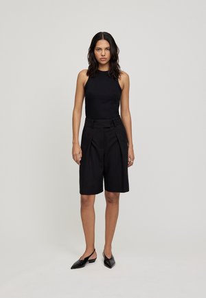 Schwarzes ärmelloses Oberteil, das mit schwarzen taillierten Shorts kombiniert wird. Die Shorts haben Falten und einen gegürteten Bund und werden mit spitzen schwarzen Ballerinas getragen.