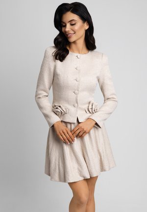 Kobieta w jasnobeżowej, teksturowanej kurtce z długim rękawem zdobionej tkaninowymi różami i pasującej krótkiej spódnicy, uśmiechająca się i patrząca w dół.