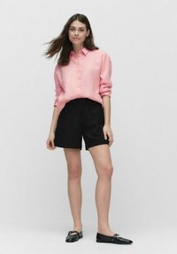 Ljust rosa linneknappskjorta, svarta shorts med elastisk midja och svarta platta skor. Modellen står på en enkel bakgrund.
