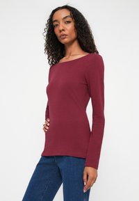 Langærmet skjorte i burgundy, lavet af et blødt stof med en bådudskæring. Sammen med blå jeans, den tætsiddende stil fremhæver formen.