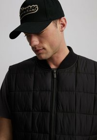 Czarna pikowana kamizelka z zamkiem błyskawicznym i okrągłym kołnierzem, noszona na ciemnym t-shircie. Czapka w kolorze czarnym z kremowym logo.