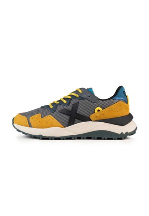 Zapatilla con parte superior de malla gris, acentos de ante amarillo y detalles en negro. Presenta una suela de goma verde y cordones amarillos brillantes.