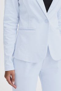 Žena u svijetloplavom uskom blejzeru s odgovarajućim hlačama i crnoj majici, ruka joj je uz tijelo s zlatnim prstenom na prstu.