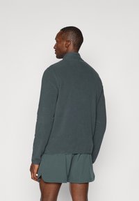 Pullover in pile verde scuro, colletto alto, maniche lunghe, texture liscia, abbinato a pantaloni corti coordinati, visto da dietro.